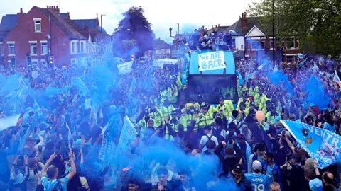 Thousands of people crowd around a bus surrounded on all sides by police officers in high-vis jackets. many of the people are carrying flares giving off lots of blue smoke. Many are also carrying light blue Coventry City flags. At the centre is the open top bus with the team at the top. At thr front is a sign, reading: "We are back"