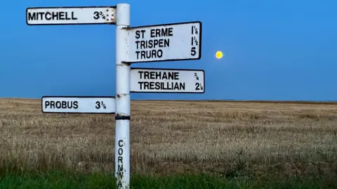 Jane Lewarne White rural signpost in St Erme, Cornwall, standing beside a harvested field at dusk, with a small golden full moon low on the horizon in the background.