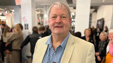 A man wearing a blue and white chequered shirt and cream suit looks into the camera.