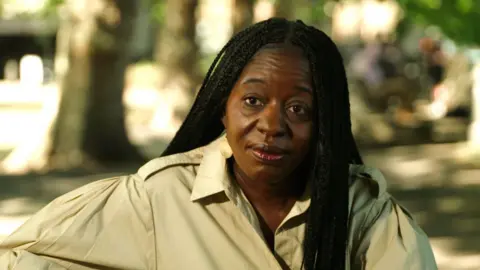 BBC A portrait photograph of a woman with black hair in braids, with blurred trees and grass in the background