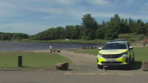 a police car is parked next to the loch. A police officer and a woman walking two dogs are on a path