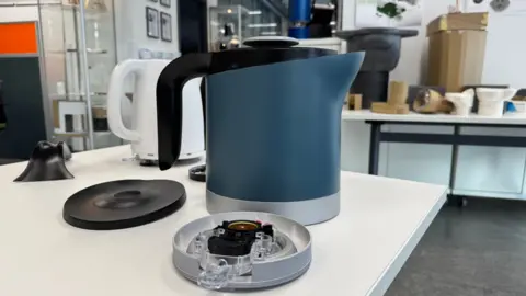 The prototype kettle in black, blue and chrome showing its base detached on a worktop, with a white kettle behind it