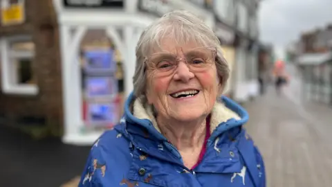 An elderly woman smiles. She is wearing a blue coat. 