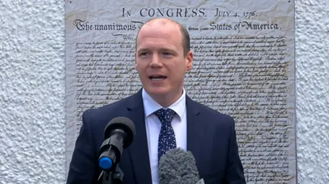Gordon Lyons is standing in front of large print of the US Declaration Of Independence. He is wearing a navy suit and has two large microphones in front of him.