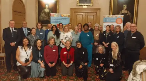 Warwickshire County Council A group of about 30 people, some kneeling and some stood, are posing for a photograph in a room with large oil paintings and a patterned carpet.