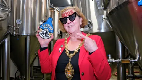 Stoke-on-Trent City Council Lord Mayor Lyn Sharpe, wearing a red jacket and gold chains of office, is holding a specially designed pump clip which promotes the centenary beer which Titanic Brewery has launched. She is also wearing heart-shaped glasses which say "2025" on the top.