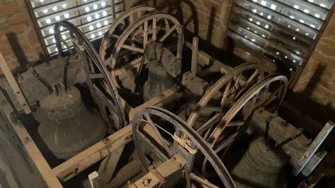 David Clements A view inside the church belfry before the restoration work was carried out. It is a small brick tower with dotted windows on each wall. On the floor there are wooden beams holding up bell frames and a wheel pulley system to ring the bells. The bells are all dirty, dark and rusty. 