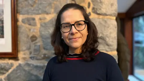 BBC A middle-aged woman in a blue jumper. She has shoulder length hair and glasses and stands by an interior granite wall with the corner of a picture frame visible.