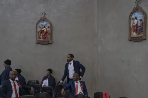 MARCO LONGARI / AFP Men dressed in matching dark suits and red tie wait in seats. Above them are depictions of the stations of the cross.