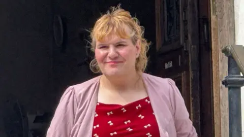 Katie Wallis walking out of court building wearing red top with white bows and pink blazer over it. She has long curly ginger hair up in a pony tail.