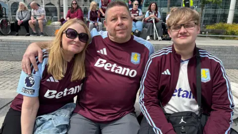 Two men and a woman wearing Aston Villa outfits sit on a low platform in Birmingham city centre.