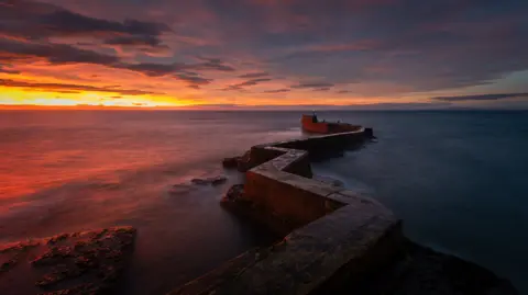 David Sneddon Landscape image of a vivid sunrise on a darkening sky, the sunset is reflecting on the water with a stone wall zig-zagging into the water and a faint green light visible in the far distance.