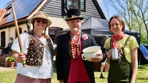 Contributed A man wearing a black top hat, black suit jacket, red T-shirt stands in between two women in a line who are smiling at the camera. He holds a white pot in one hand. The woman to his left wears a cream hat, sunglasses, white blouse, waistcoat with flowers on it and is holding a stick in her left hand. The other woman has short brown hair and is wearing a green T-shirt with green dungarees. She is holding a metal tankard in one hand.