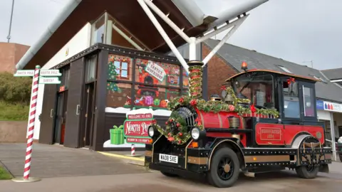 East Riding of Yorkshire Council Bridlington's land train parked up at a station, it is red and black, with gold trim and decorated in tinsel and baubles. A red and white pole pointing left to the south pole and right to the north pole is in front of the train to create a festive feel