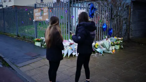 Two girls standing in front of a vigil for Max and Mason, where flowers are laid against a railing and a stop knife crime banner