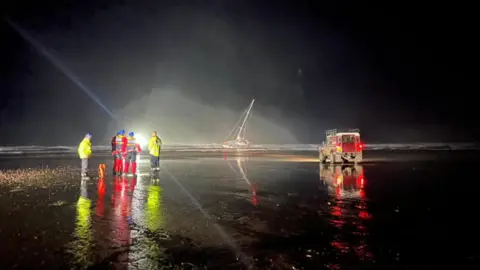 Four rescue service operatives in fluoro jackets of red and yellow look out to sea where a single-masted yacht is lying beached on the sand. A red Land Rover with its lights on in the dark is on the beach.