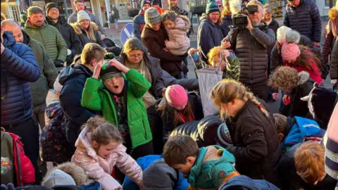 Children in the street in Driffield wearing winter coats and hats. A crowd of adults, including a man with a camera, watch on as the youngsters pick up sweets