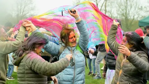 Amith Nair Three women dancing with a colourful scarf among a crowd. They have various colours of powder on them.
