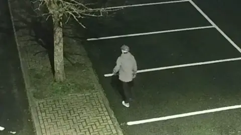 North Yorkshire Police A CCTV image of a man walking through a car park wearing dark trousers, a grey jacket, trainers and a hat.