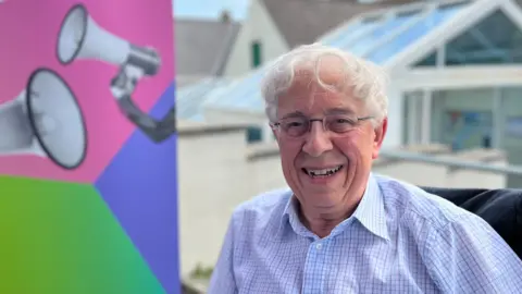 BBC A man with white hair and glasses, wearing a blue and white checked shirt. On the left of the picture is the Your Voice, Your Vote logo.
