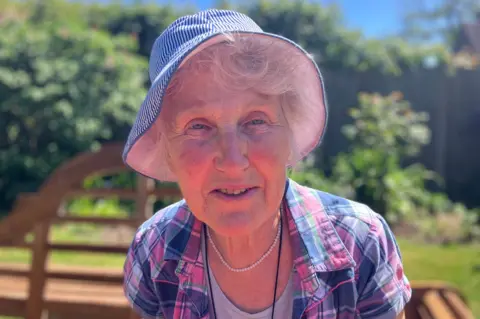 Woman is hat with pink and purple checked shirt with white necklace
