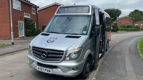 Image of the number 65 bus. It is a silver Mercedes minibus parked up at the side of a residential road.