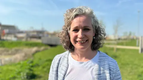 Rebecca Britton smiling into camera with green space and a housing development in the background. She has curly grey hair and is wearing a grey cardigan and white t-shirt.