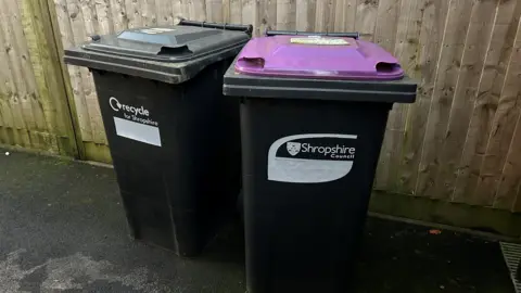 Two black bins in front of a wooden fence - one of them has a black lid, and the other has a purple lid. Both have the words Shropshire Council on the side of the bins in white lettering.