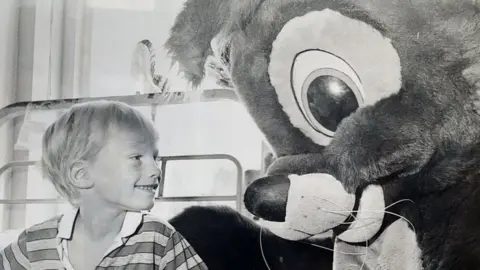 Colin in hospital bed with a mascot visiting