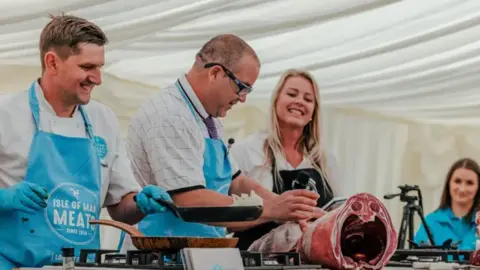 IOM GOVERNMENT Image of a previous year's festival. Representatives of Isle of Man Meats are standing on a stage in a marquee showing their product to people.
