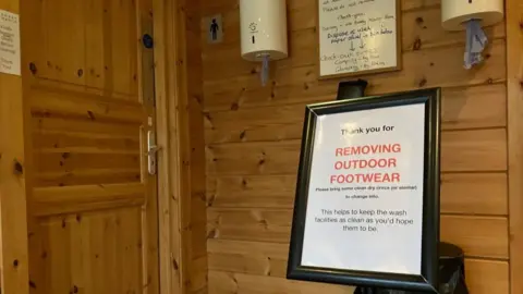 BBC/Seb Cheer A sign on a stand which says "Thank you for removing outdoor footwear. Please bring some clean dry crocs (or similar) to change into." In the background is a door with the gents logo next to it.