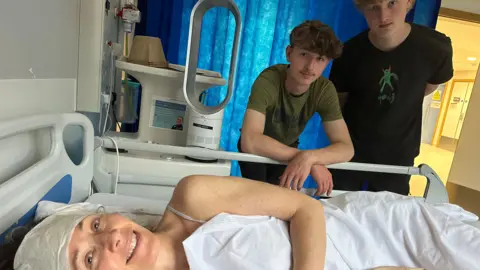 Family photo Dr Hard smiling while recovering in a hospital bed in London with her sons Callum and Oliver by her side.