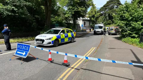 A police cordon blocking access to Clifton Suspension Bridge. A police officer stands with her back to the camera in front of a blue and white striped line of tape and a sign reading 'police' in the road. A number of police vehicles can be seen in the area.