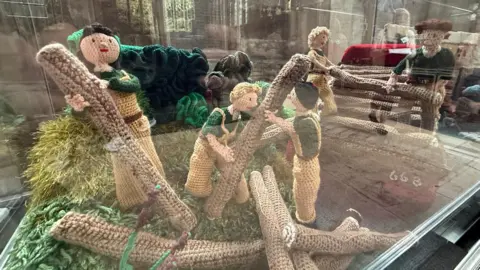 Peterborough Cathedral A display of knitted figures, shot through glass, showing the work of the Women's Land Army. Three female figures in light brown overalls are moving logs.