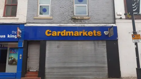 The shop on Blandford Street. It is a two-storey building with a tatty shutter over the bottom floor and a yellow sign reading "Cardmarkets". There are two windows upstairs.