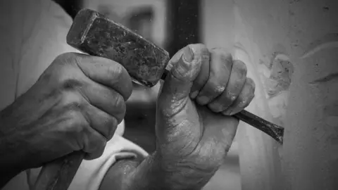 LANSS A stock black and white close-up image of the hands of a stonemason working a block of stone with a hammer and chisel. 