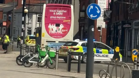 BBC Police vehicles with flashing lights parked behind a bus at a town centre junction, with part of the road cordoned off, bicycles parked nearby and surrounding shops and buildings visible