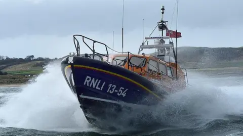 The blue and orange RNLI lifeboat whips up waves at it travels through the sea.