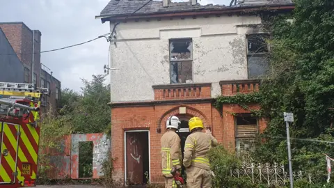 A badly damaged building which has a section of roof missing and smashed windows. There are two firefighters stood in front of the building wearing hard hats and fire suits. 