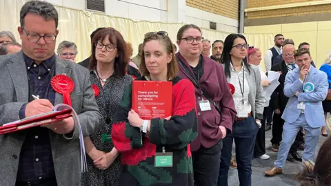 NicolaHaseler/BBC A group of people stand around clustered together waiting for results. In the foreground are Labour candidates and supporters wearing red rosettes, while on the right back side of the photo are Reform teams in blue.