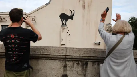 EPA A man and a woman with smartphones in their hands taking pictures of piece of art by street artist Banksy. The work itself is of a goat and it is on the side of a building in Kew in south-west London. 