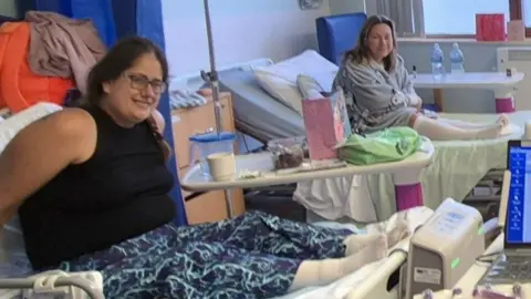 Two women in hospital beds smiling for the camera. On the left is a woman with brown hair in a plait and glasses, who is leaning forward on a bed, wearing a black vest and blue patterned trousers. Next to her is another woman in a grey jumper and knee high socks with long blonde hair leaning forward and smiling too.