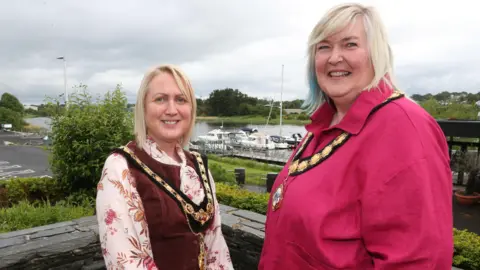 Causeway Coast & Glens Borough Council Dawn Huggins and Sandra Hunter are standing outside with bushes behind them. Boats can be seen in a lough behind them. Both are wearing gold chains of offices. Huggins is wearing a floral top and a maroon dress and Hunter is wearing a pink shirt. 