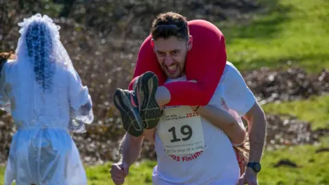 Getty Images A man carrying a woman on his back running up a hill. A person in a wedding dress is in the background.