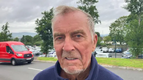 A man in a blue jacket and with short grey hair, stands in front of a car park