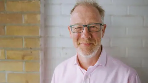 Ben wearing a pastel pink shirt with ginger and grey stubble wearing blue rimmed glasses smiling at the camera. He is standing in front of a brick wall. Some of it has been painted white.