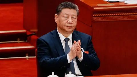 Chinese President Xi Jinping adjusts his navy blue jacket as he sits at the closing session of the Chinese People's Political Consultative Conference (CPPCC) at the Great Hall of the People on March 11, 2026 in Beijing, China. He is sitting in front of a long wooded table with two white tea cups in front of him.