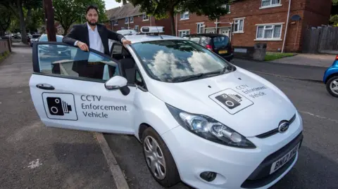 A man in a black suit and white shirt standing at the door of a white Ford in a residential street. One hand is resting on the car roof, the other on the open door. The car has CCTV Enforcement Vehicle written on the front and door, along with an image of a camera. Parked cars and semi-detached homes can be seen in the street behind.