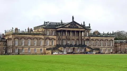 An image of Wentworth Woodhouse a large stately home. The front facade   is about 600ft long. It is is a symmetrical structure with a central building, two-story flanks, three-story porticoed wings, and small towers at each end.  