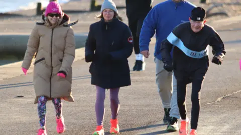 John Nurden Elderly man in black hat running alongside two women in coats and hats